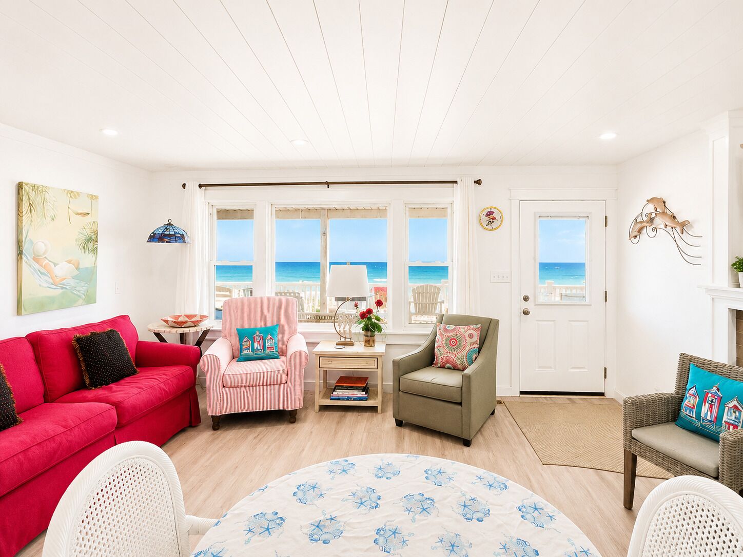 A living room with a red sofa, a striped armchair, a round table, and a view of the ocean through large windows.