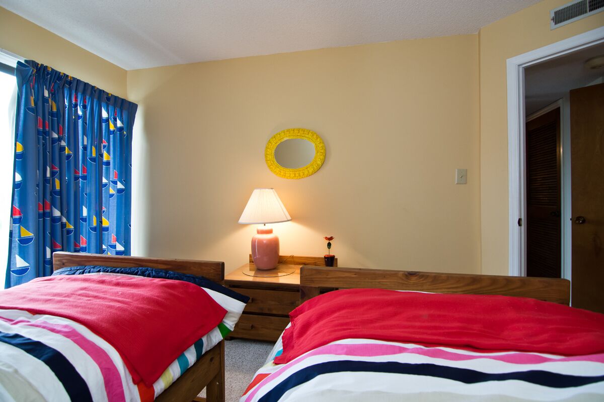 Une chambre avec deux lits, une table de chevet avec une lampe, un miroir au mur et une porte sur le côté droit.