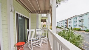 A porch with white rocking chairs and a red table, overlooking a street with multi-story buildings. - Pebble Beach E-102, Oceanview Condo in Emerald Isle, NC! (Emerald Isle)