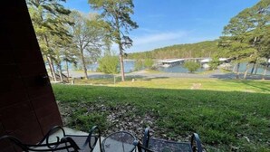A patio with two metal chairs and a small round table, overlooking a grassy area with trees and a body of water. - Mountain Harbor Queen Guest Room on Lake Ouachita by RedAwning (Mount Ida)