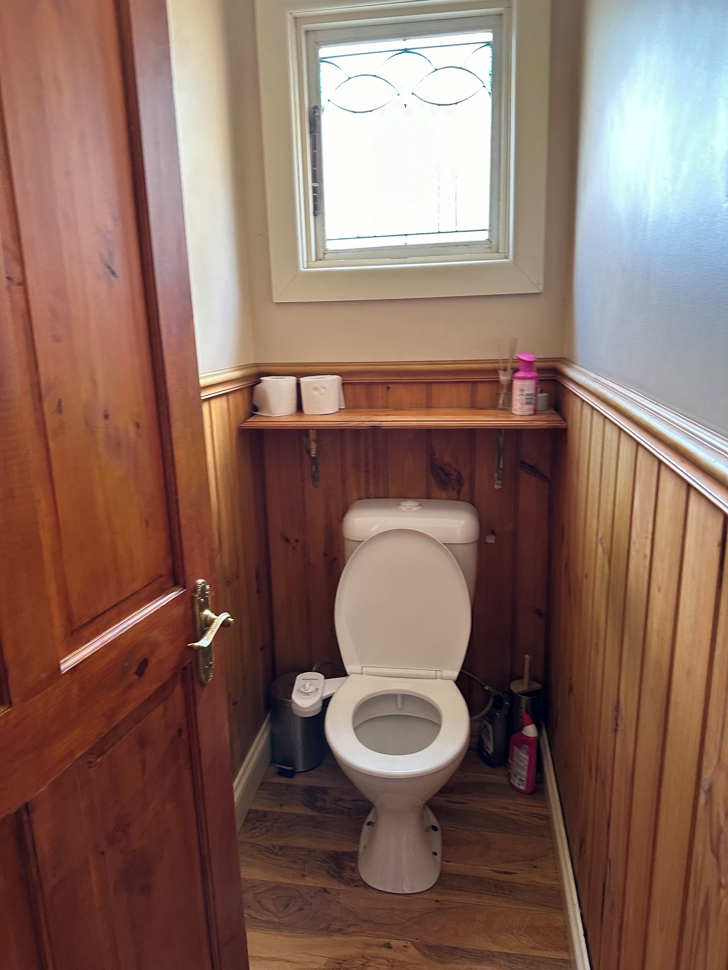 Ein kleines Badezimmer mit Holzboden, Toilette in Weiß, einem Fenster über der Toilette und einem Ablagefach für Pflegeartikel.