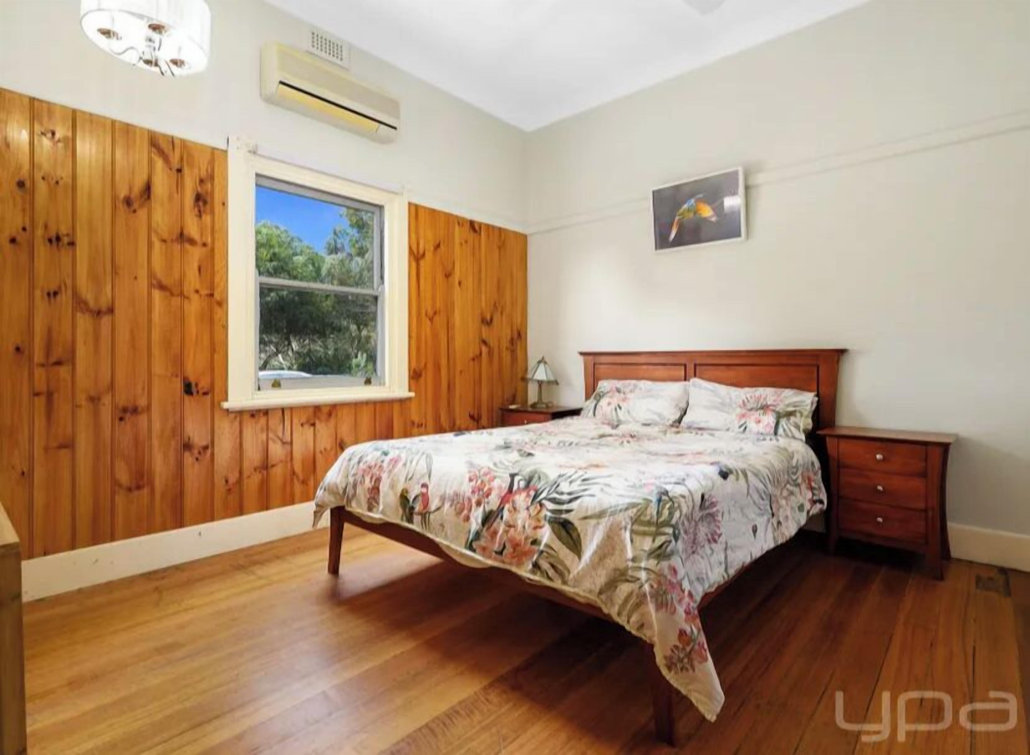 Een slaapkamer met een houten lambrisering, een groot raam, een bed met bloemenbeddels, een nachtkastje en een aan de muur bevestigde airconditioner.