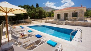 Uma piscina com espreguiçadeiras e um guarda-sol, um edifício de pedra com telhado em telhas, e montanhas ao fundo.