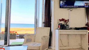Ein Zimmer mit großem Fenster mit Blick auf einen Strand, ein Korbstuhl, ein kleiner Tisch und ein Fernseher auf einem Schrank.