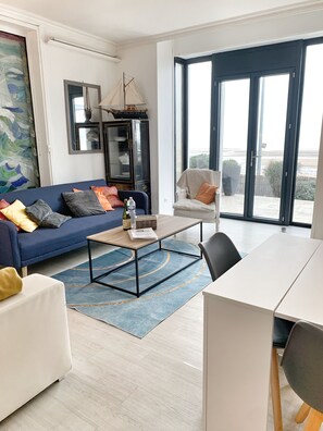 A modern living room with a sofa, a coffee table, a dining table, and a chair. There is a large window with a view of the beach.