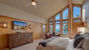 A bedroom with a large bed, a wooden dresser, a TV, and a view of the outdoors through large windows. - Ramblin' Rose - Lakeview - TLUXP (Tahoe Vista)