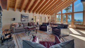 A spacious living room with a high wooden ceiling, large windows offering a view of the outdoors, and a cozy seating area with a fireplace. - Ramblin' Rose - Lakeview - TLUXP (Tahoe Vista)
