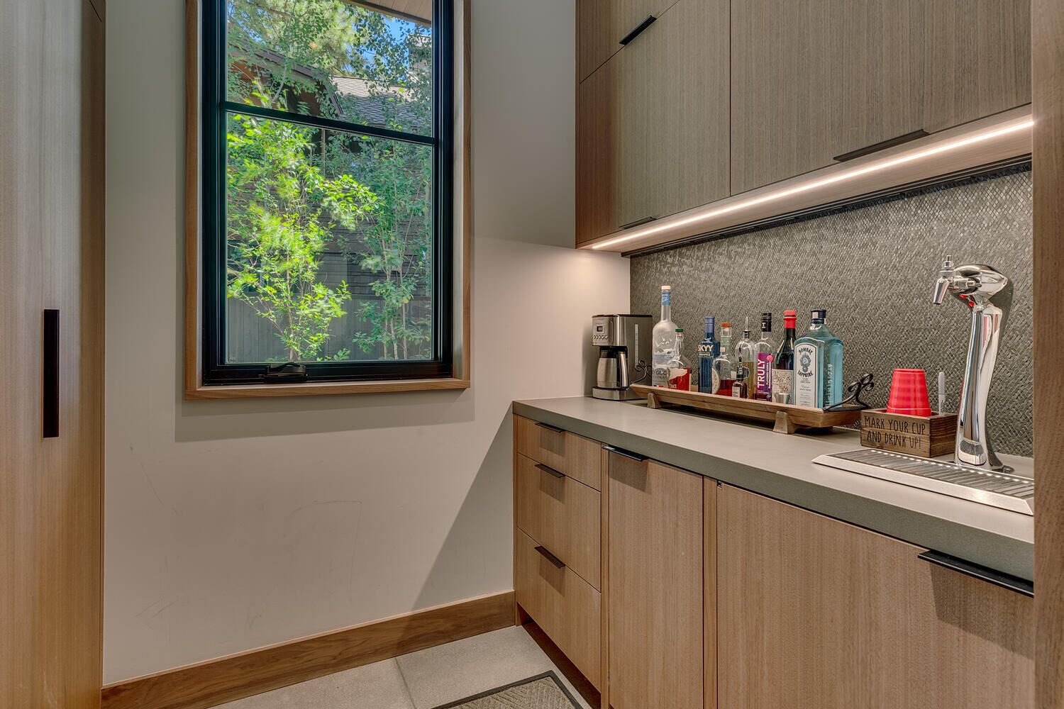 Een moderne keuken met een groot raam, houten kastjes en een aanrecht met verschillende flessen en een koffiezetapparaat.