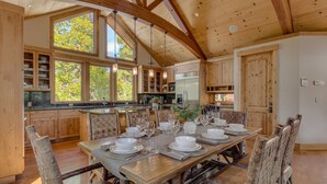 Una cocina amplia con armarios de madera, un comedor preparado para seis personas con una mesa larga, y ventanas grandes que dejan entrar luz natural.
