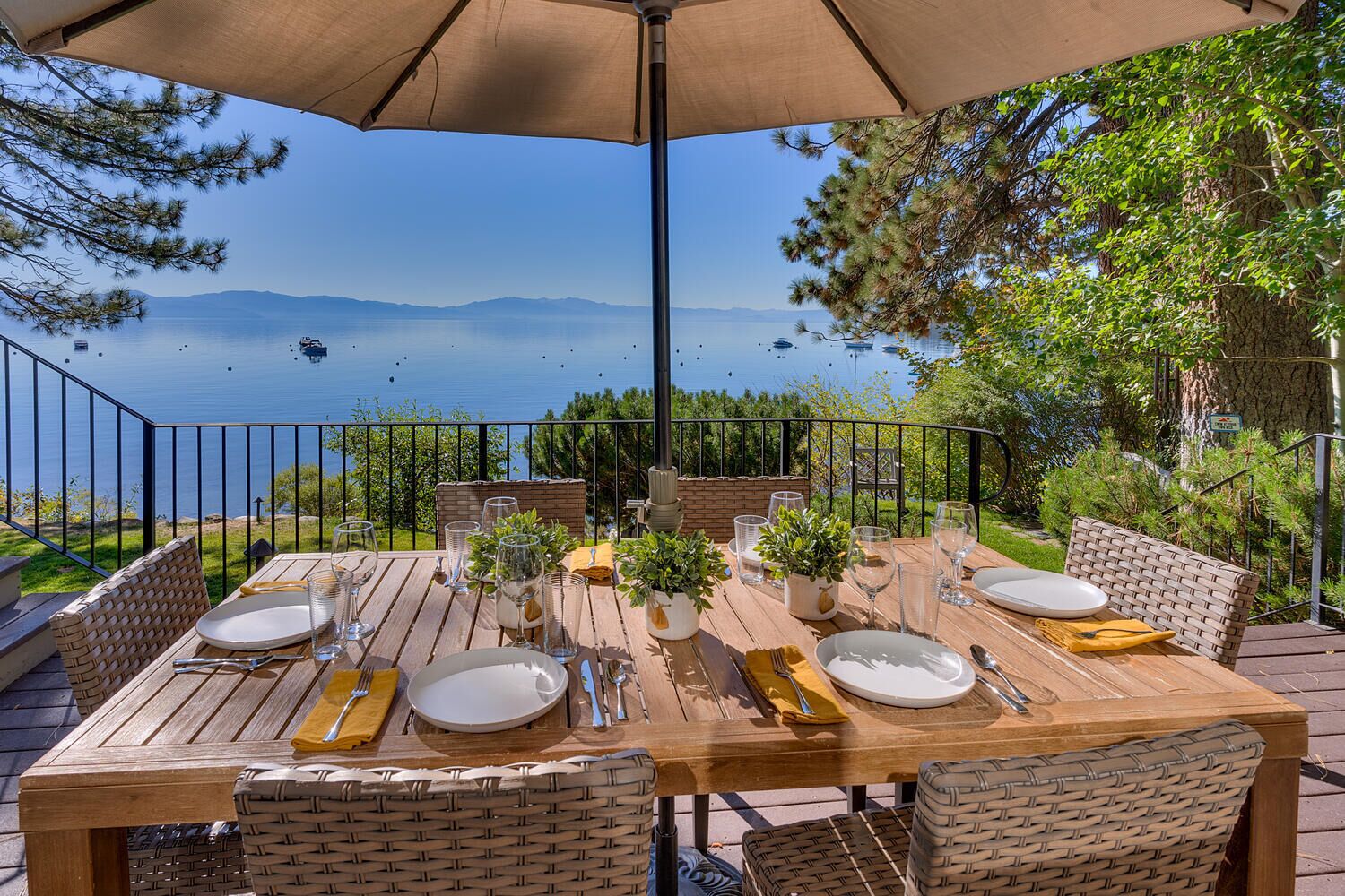 Un espace de restauration en plein air avec une table dressée pour six personnes, offrant une vue sur un lac où l’on aperçoit des bateaux et, au loin, des montagnes.
