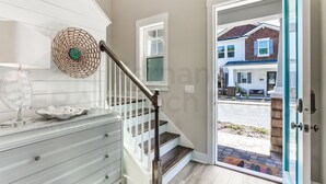 A staircase with a wooden handrail, a white wall with a decorative woven item, and a view of a residential street through an open door.
