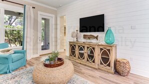 A living room with a teal sofa, a round wicker ottoman, a wooden cabinet with glass doors, and a flat-screen TV mounted on the wall.
