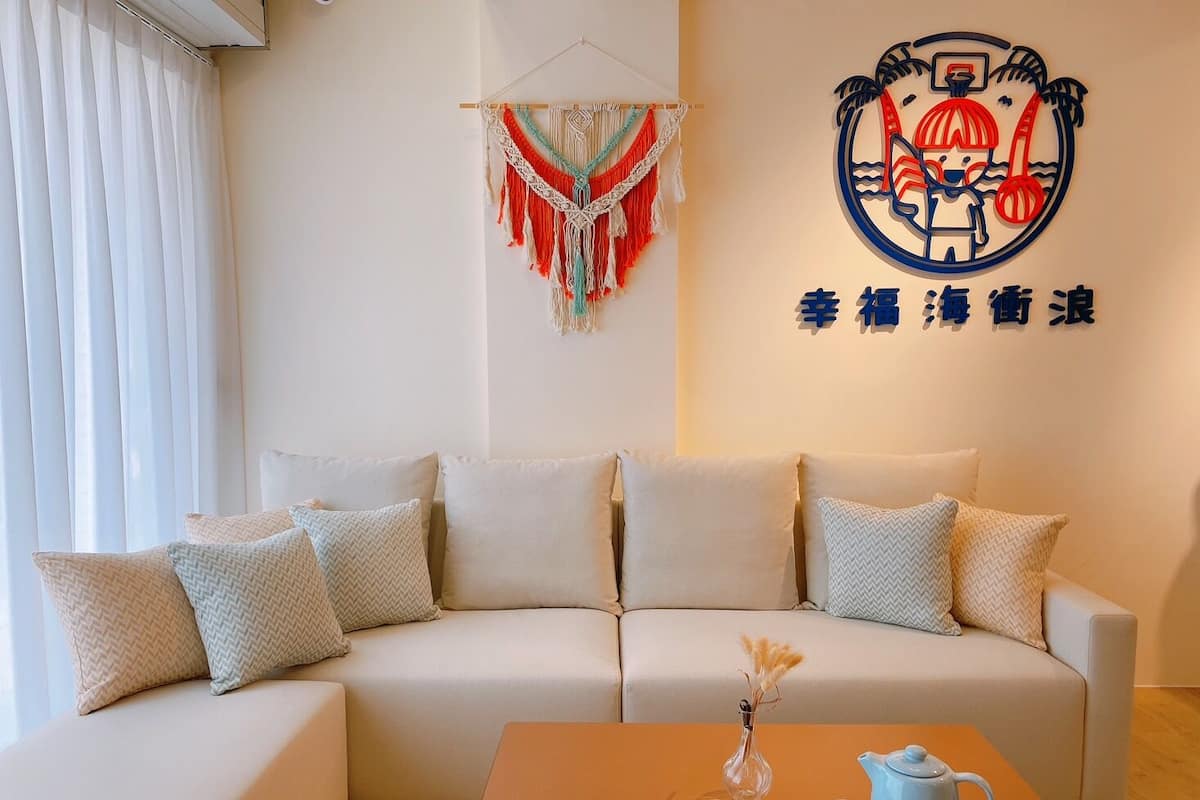 A living room with a sofa, pillows, a macrame wall hanging, and a wall with a logo and text.