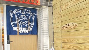 Una entrada de hotel con una puerta de madera, un cartel azul con una palmera y el texto 'HAPPY SEA', y un felpudo que dice 'HELLO YOU LOVELY PEOPLE'.