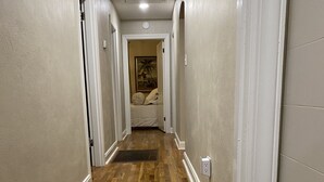 Un couloir doté d’un plancher en bois, de murs blancs et d’une porte donnant sur une chambre à coucher où se trouvent un lit et un tableau.
