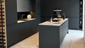 Eine moderne Küche mit dunklen Schränken, einer zentralen Kochinsel und einem Weinkühlschrank auf der linken Seite.
