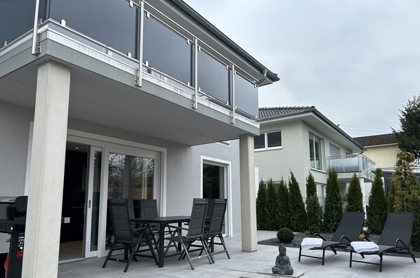 Una casa moderna con un porche cubierto, un comedor con mesa y sillas, y un área de descanso junto a la piscina.