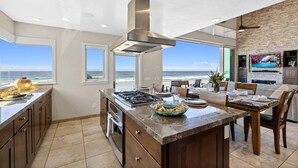 A modern kitchen with a large island, a gas stove, and a stainless steel range hood. There are large windows offering a view of the ocean. - Cielo Mar | 4BR Oceanfront Escape + Rooftop (San Diego)