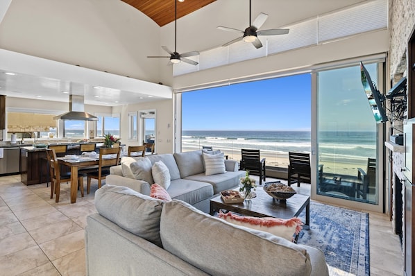 A spacious living room with a large window overlooking the ocean, a kitchen in the background, and a ceiling fan. - Cielo Mar | 4BR Oceanfront Escape + Rooftop (San Diego)