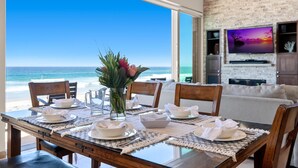 A dining table set with plates, bowls, and a vase of flowers, overlooking a beach with a clear blue sky. - Cielo Mar | 4BR Oceanfront Escape + Rooftop (San Diego)