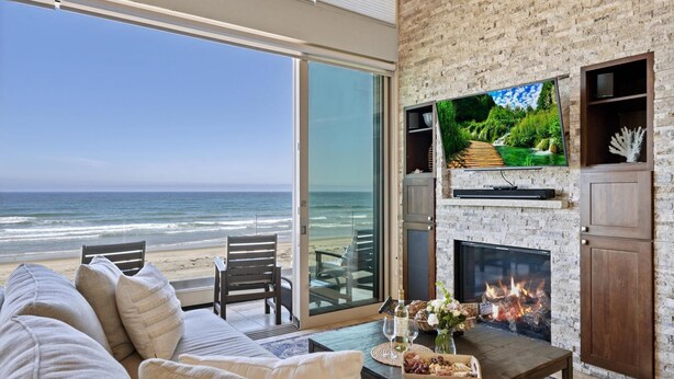 A living room with a large TV, fireplace, and a view of the beach. - Cielo Mar | 4BR Oceanfront Escape + Rooftop (San Diego)