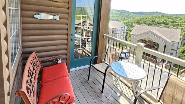 A wooden deck with a red cushioned bench, a small table, and a chair. A teal door with a window is visible, and a fish decoration hangs on the wall.