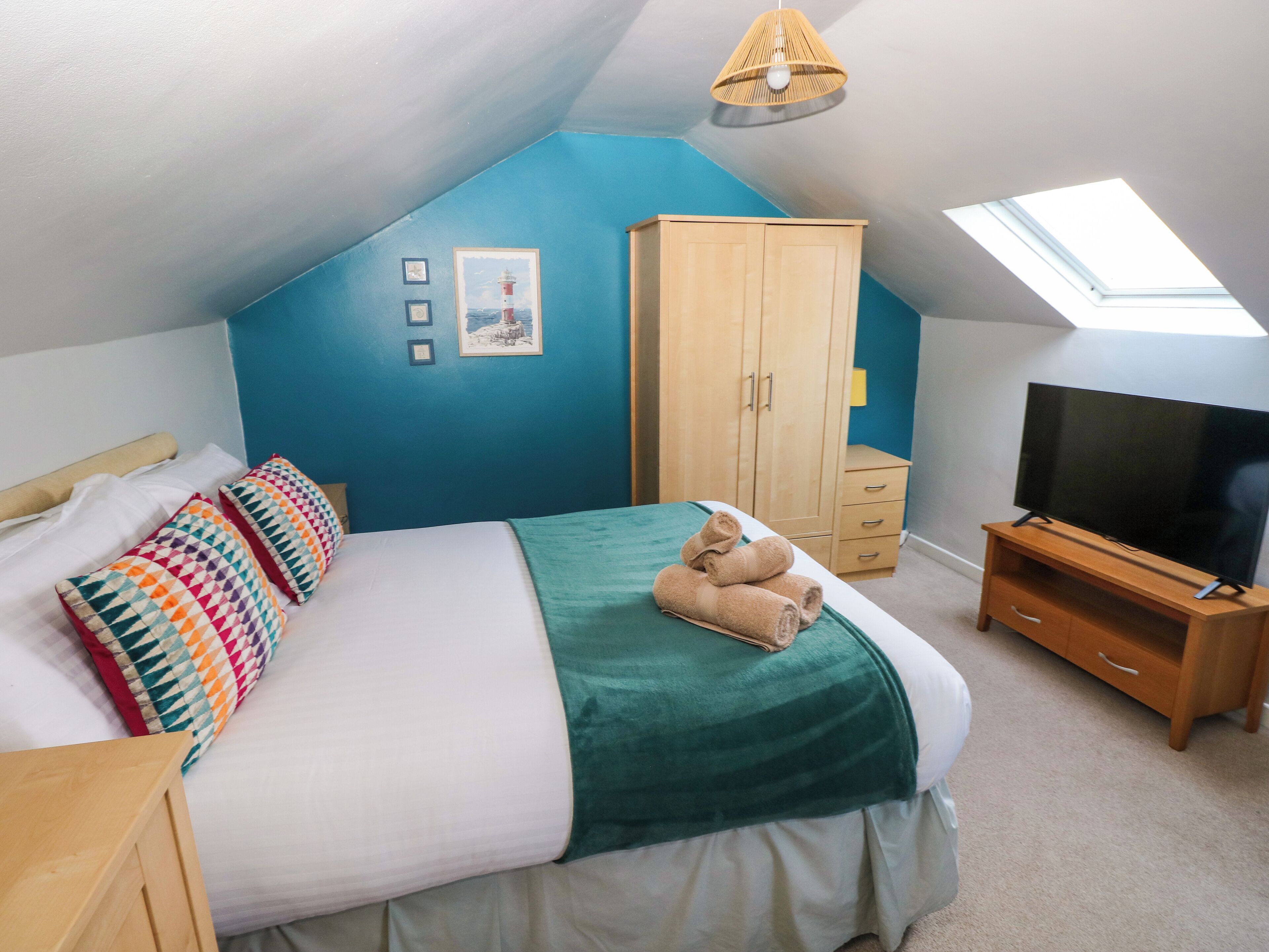 Une chambre avec un plafond en pente, un lit agrémenté de coussins colorés, une couverture verte, une armoire en bois, un téléviseur posé sur un meuble en bois et une lucarne.
