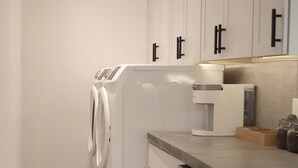 Une cuisine compacte équipée d’un lave-linge et d’un sèche-linge intégrés, d’un lave-vaisselle en acier inoxydable et d’un plan de travail sur lequel se trouve une machine à café.