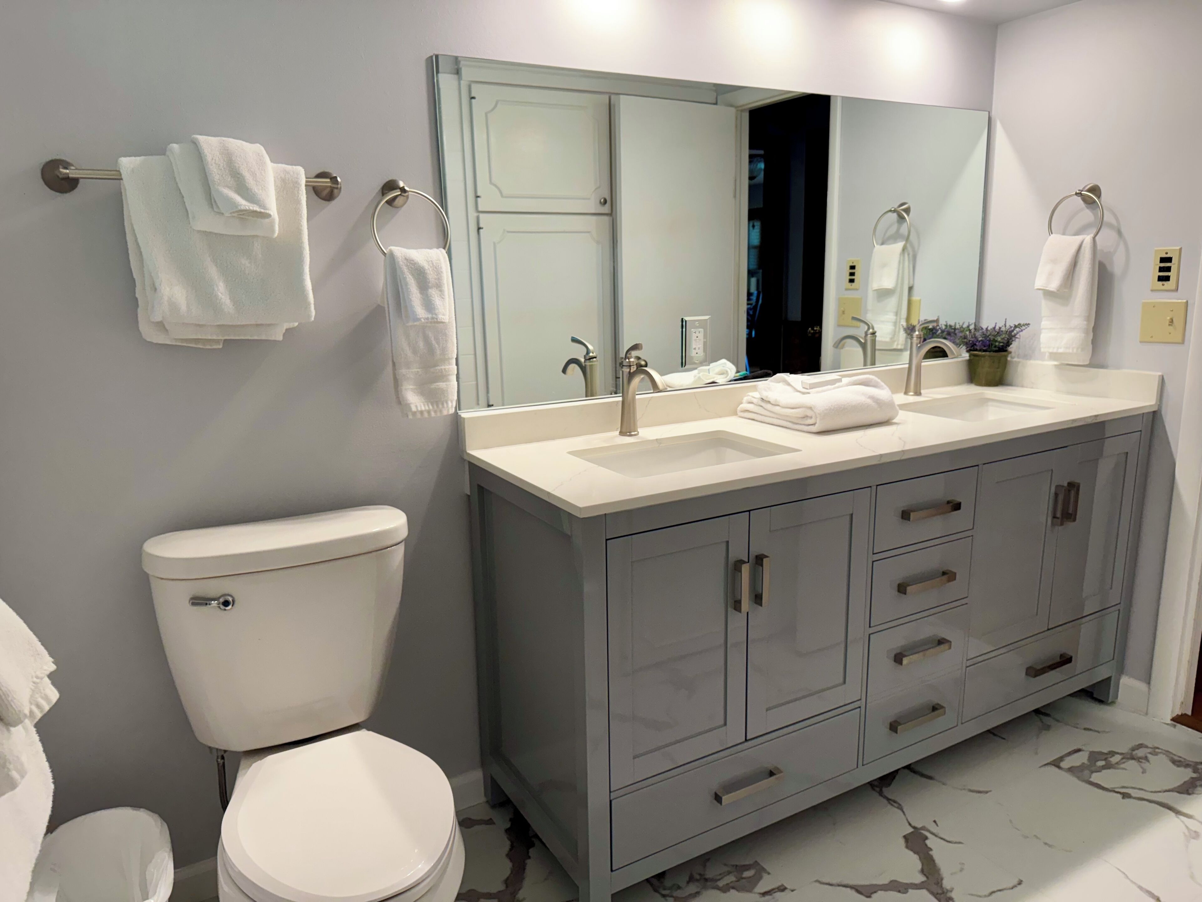 A modern bathroom with a white toilet, a double sink vanity with a large mirror, and a grey cabinet with drawers.