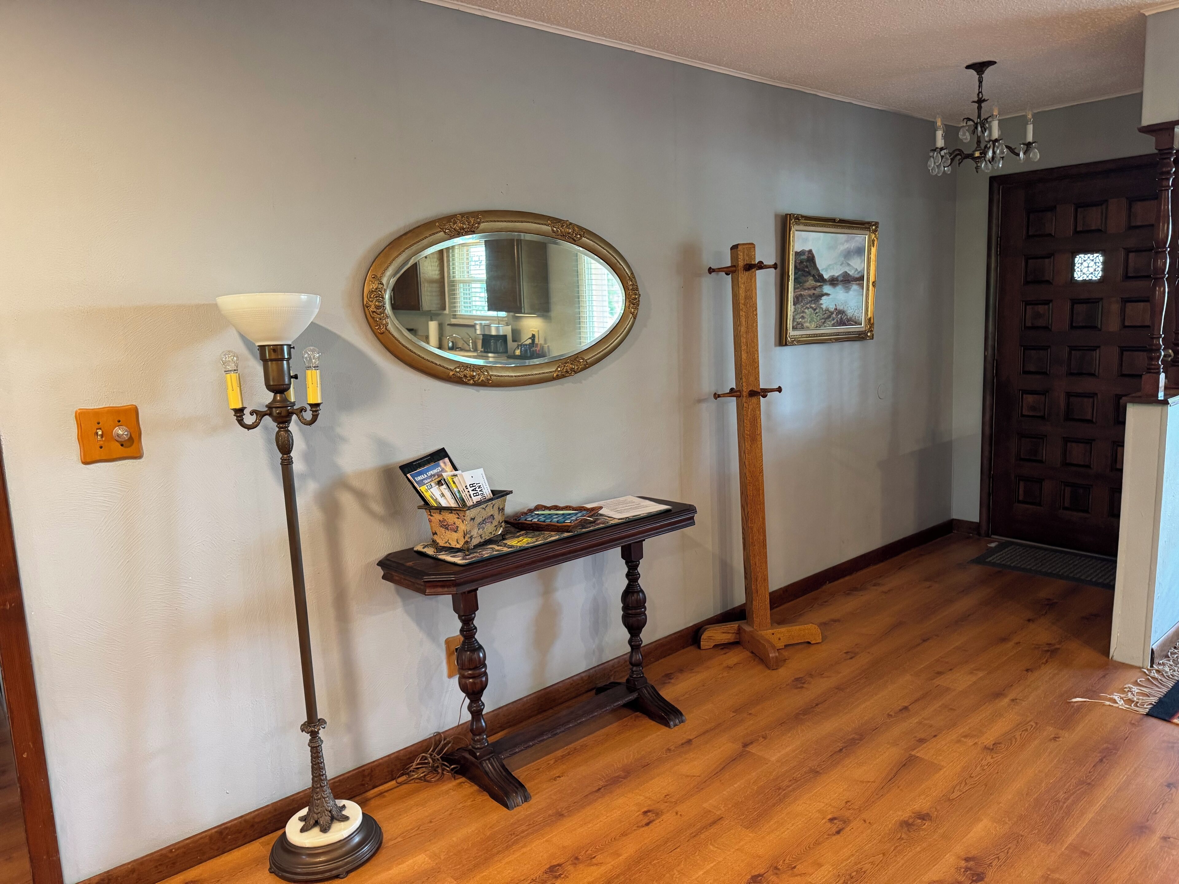 A hallway with a wooden floor, a wall-mounted mirror, a table with magazines, a floor lamp, and a door with a decorative panel.