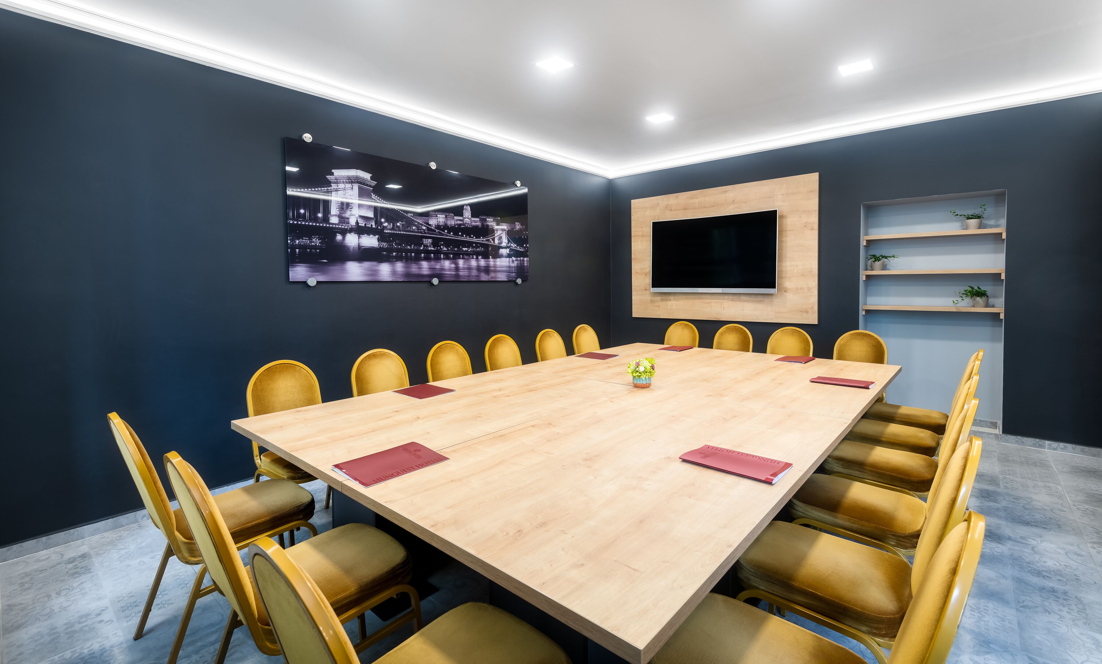 Una sala de conferencias con una gran mesa, sillas amarillas, un televisor montado y un cuadro de un paisaje urbano en la pared.