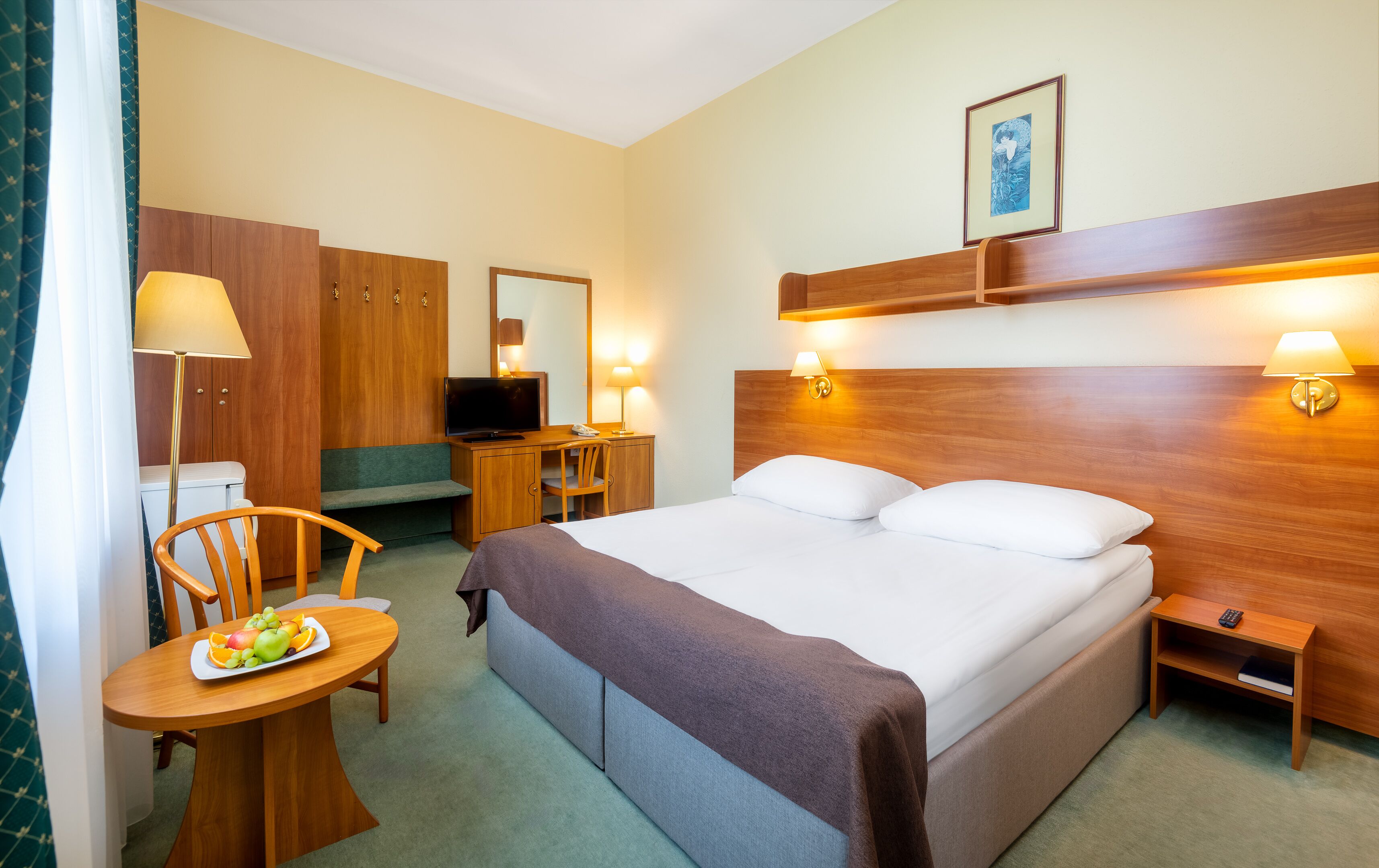 A hotel room with a bed, bedside table, lamp, desk, chair, and a small table with fruit.