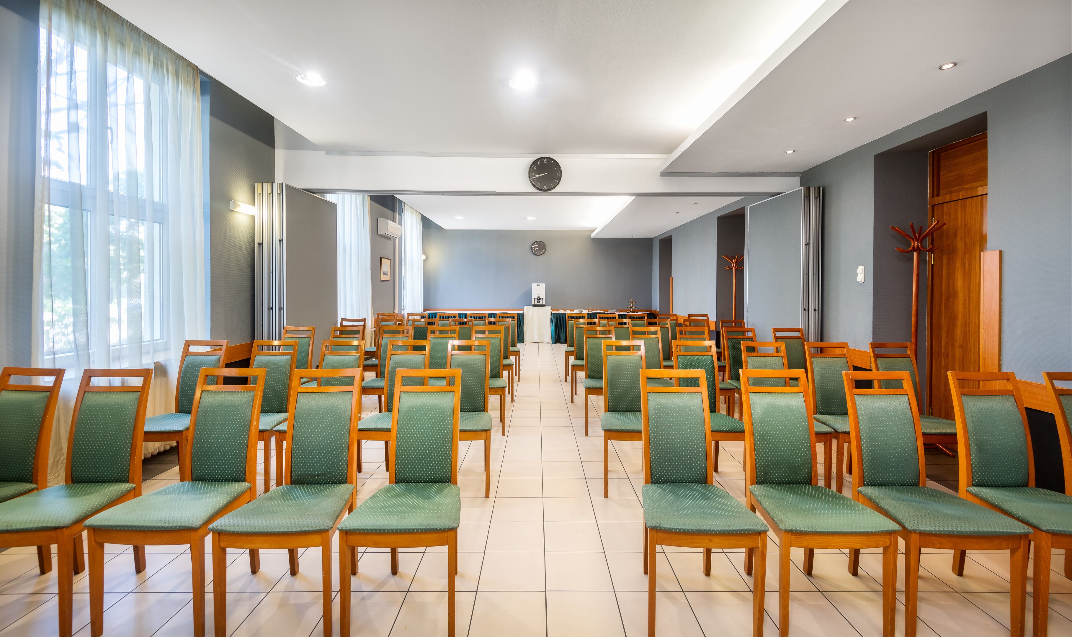 A conference room with rows of wooden chairs and green cushions, a clock on the wall, and a water dispenser in the center.