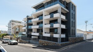 Um edifício residencial moderno, com vários andares, varandas, fachada de pedra e área de estacionamento com carros.