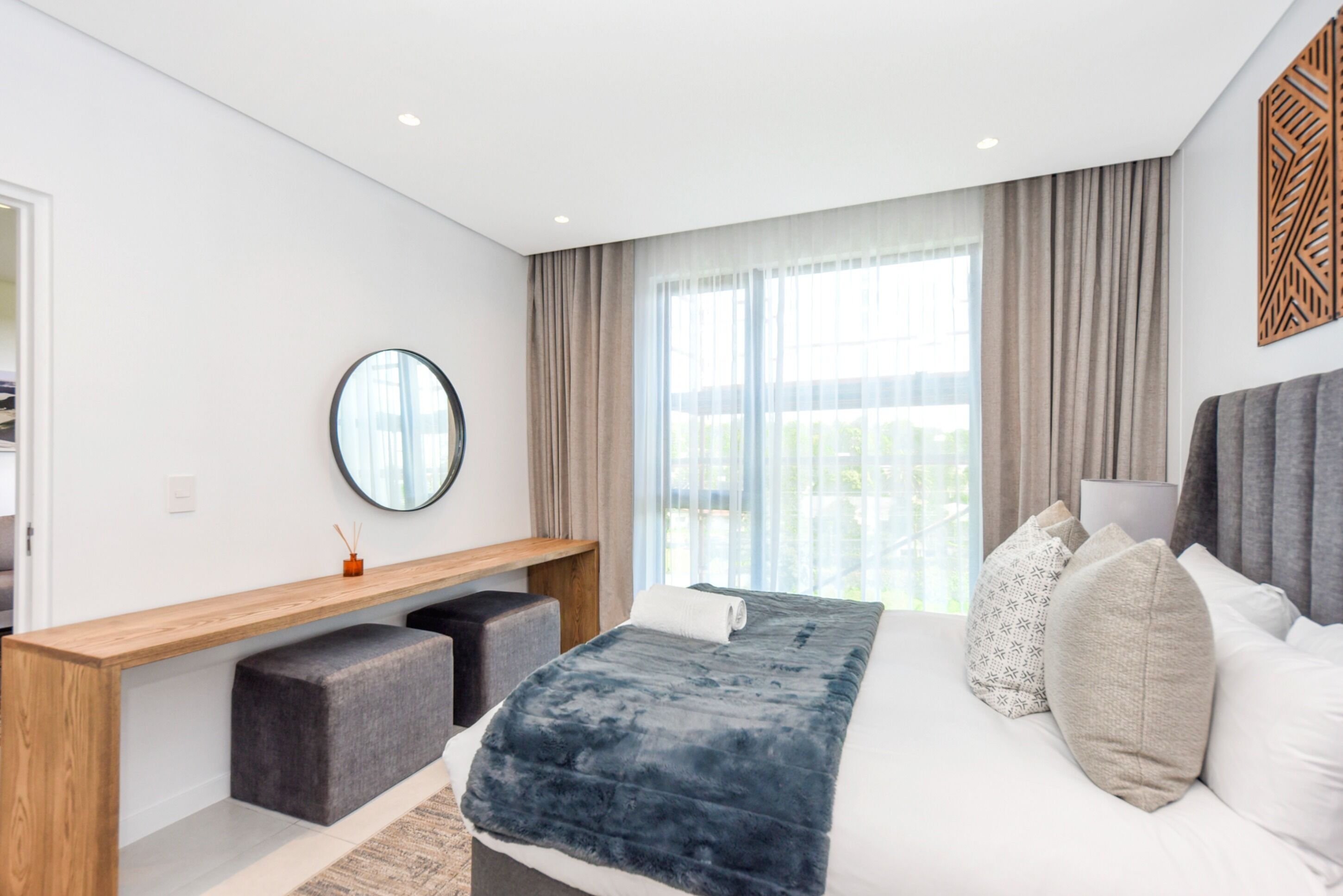 A modern hotel room with a large bed, a wooden console table, a round mirror, and a view of the outdoors through large windows.