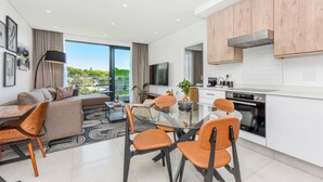 A modern living room with a sofa, dining table, and chairs, a kitchen with white cabinets and appliances, and a large window offering a view of greenery. - Luxury 2 Bed in Sandton Gate (Johannesburg)