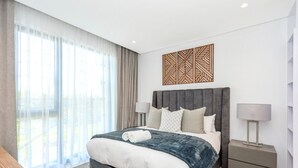 A modern hotel room with a large bed, two bedside tables, a decorative wall piece, and a view through large windows. - Luxury 2 Bed in Sandton Gate (Johannesburg)