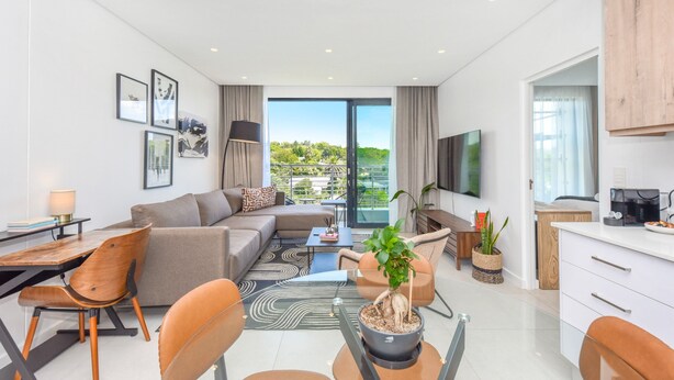 A modern living room with a sofa, coffee table, and dining area. There is a large window with a view of greenery. - Luxury 2 Bed in Sandton Gate (Johannesburg)