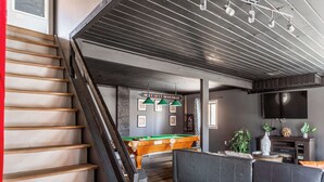 A staircase, a pool table, a sofa, and a television in a room with a wooden floor and gray walls.