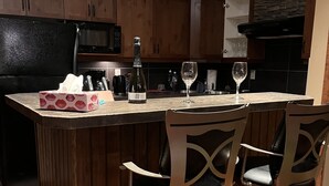 A modern kitchen with wooden cabinets, a black refrigerator, a microwave, and a wine bottle on the island. There are three bar stools with cushioned seats. - 4-Bedroom Condo in Radium, BC (Radium Hot Springs)
