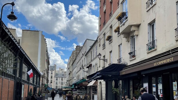 Une rue pavée bordée de cafés et de restaurants, avec des terrasses et un ciel d’un bleu éclatant.