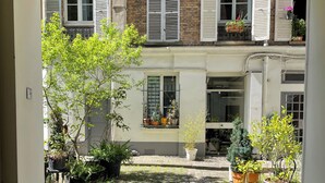 Une cour avec un chemin pavé, des plantes en pot et un bâtiment aux fenêtres fermées par des volets.
