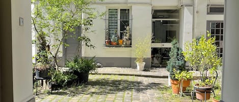 A courtyard with a cobblestone path, potted plants, and a building with shuttered windows.