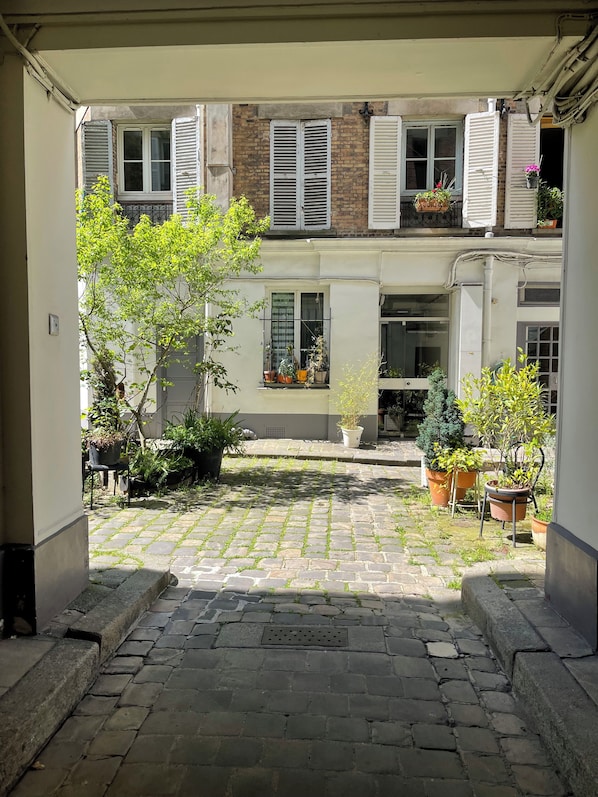 A courtyard with a cobblestone path, potted plants, and a building with shuttered windows.
