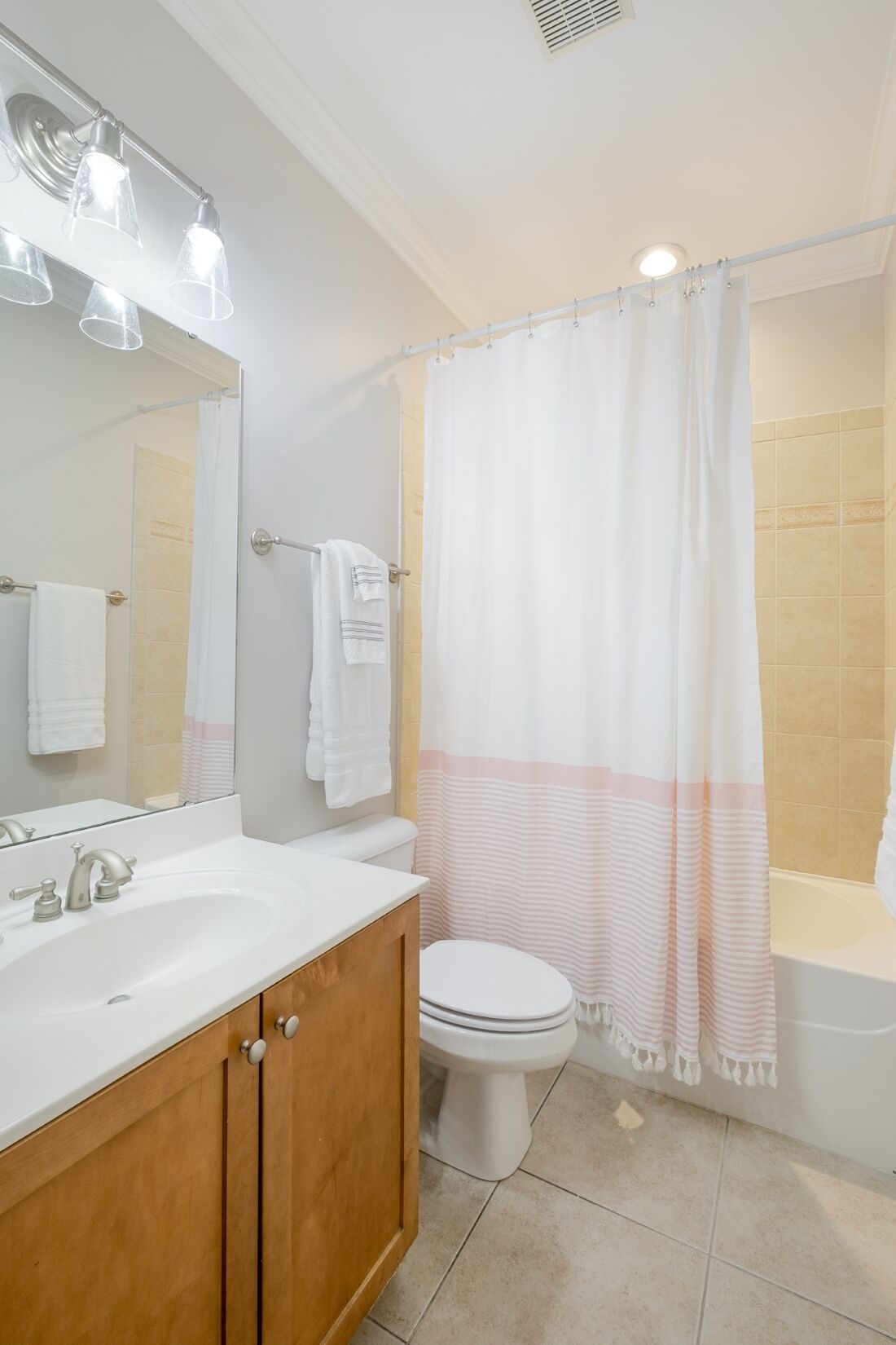 A bathroom with a white sink, wooden vanity, a white toilet, a shower with a striped curtain, and a mirror with lights.