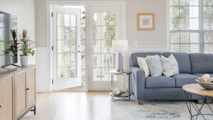 A living room with a grey sofa, a small round table with a lamp, a television on a cabinet, and a door leading outside. - Pie Seas - 4 bedroom with elevator, dog friendly! (Carolina Beach)
