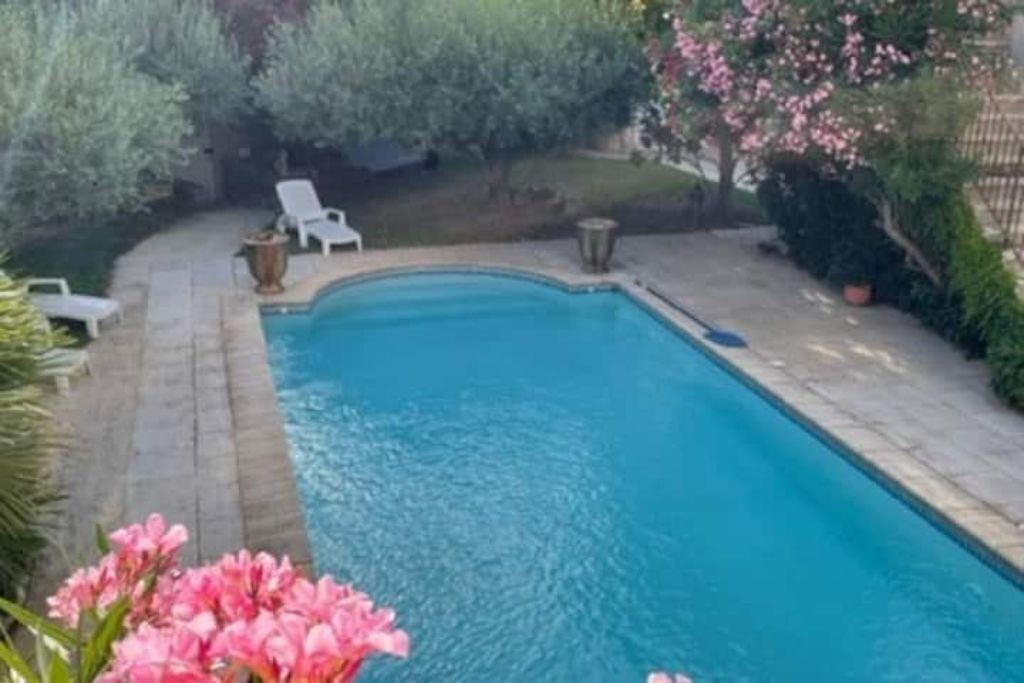 A rectangular swimming pool with a stone deck, surrounded by greenery and a few white lounge chairs.