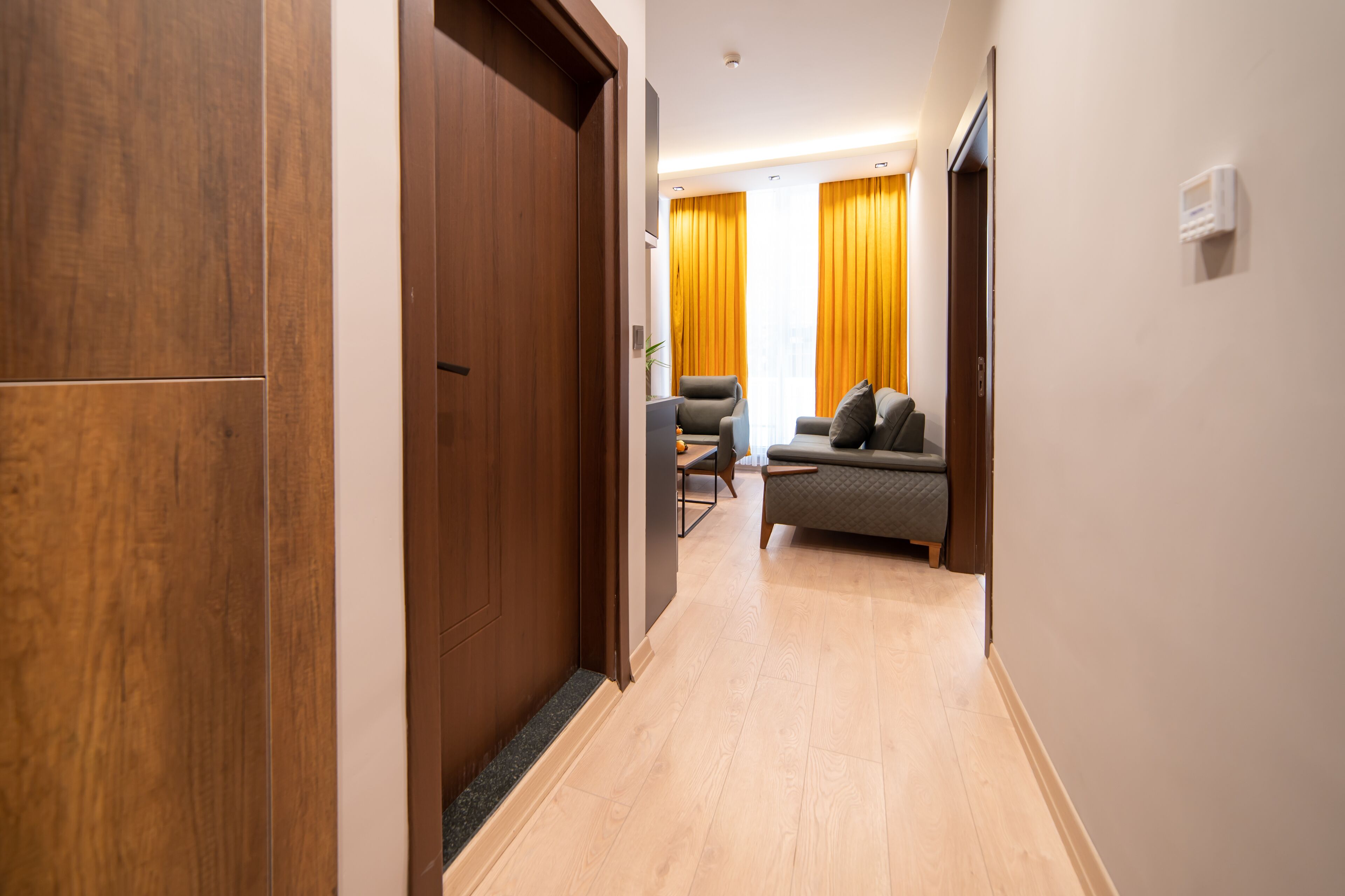 A hallway with a wooden door, a sofa, and a chair in a modern room with light wooden flooring and yellow curtains.