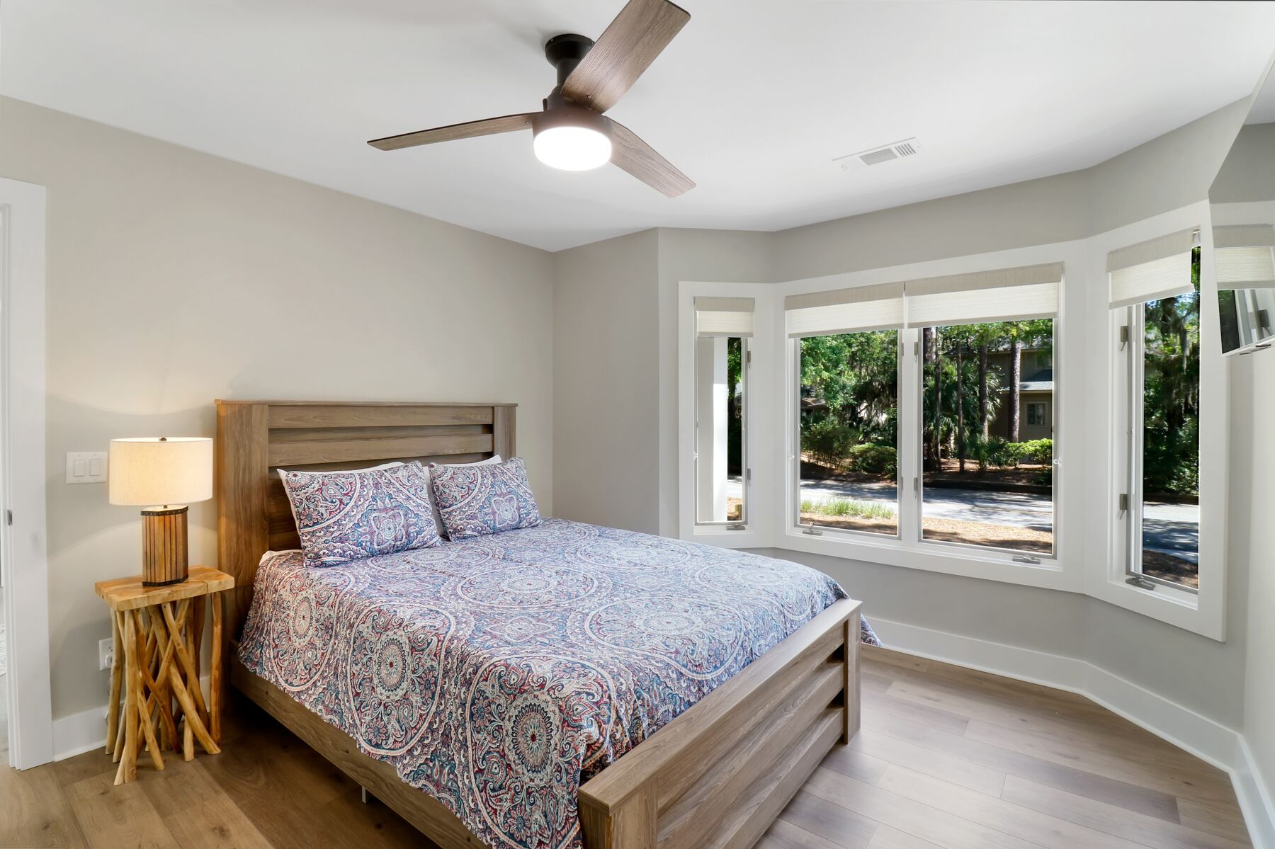 A bedroom with a bed, a standing lamp, a ceiling fan, and large windows offering a view of the outdoors.