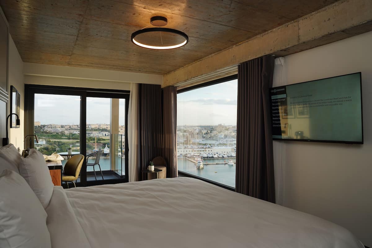 Habitación de hotel con una cama grande, un escritorio y una silla. Se ve un paisaje urbano y una masa de agua a través de amplias ventanas.